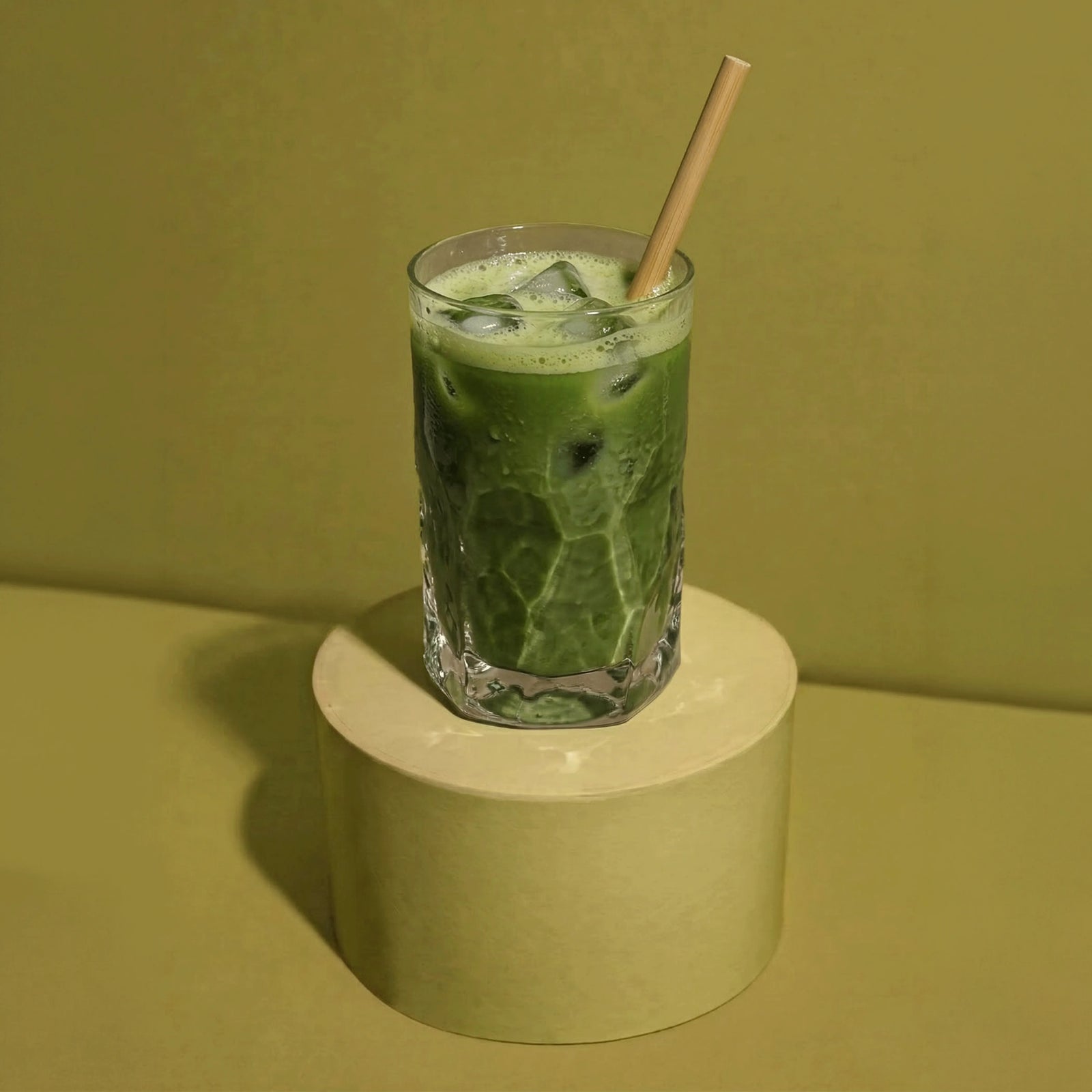 Erfrischender Iced Matcha Latte mit Eiswürfeln und einem Bambusstrohhalm in einem transparenten Glas, stehend auf einem grünen Podest.