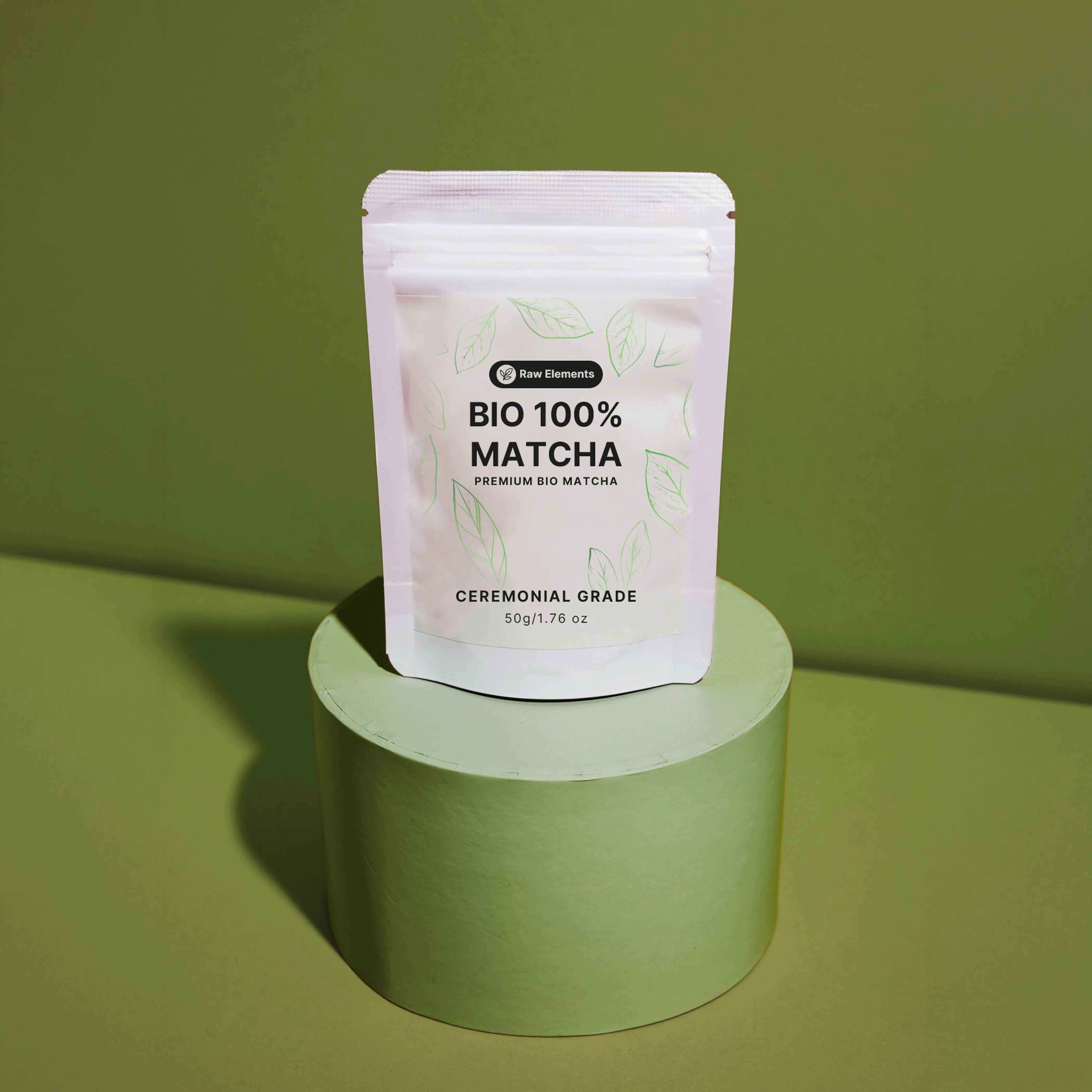 Weißer Standbodenbeutel des Raw Elements Bio 100% Matcha Ceremonial Grade, platziert auf einem runden, olivgrünen Podest.