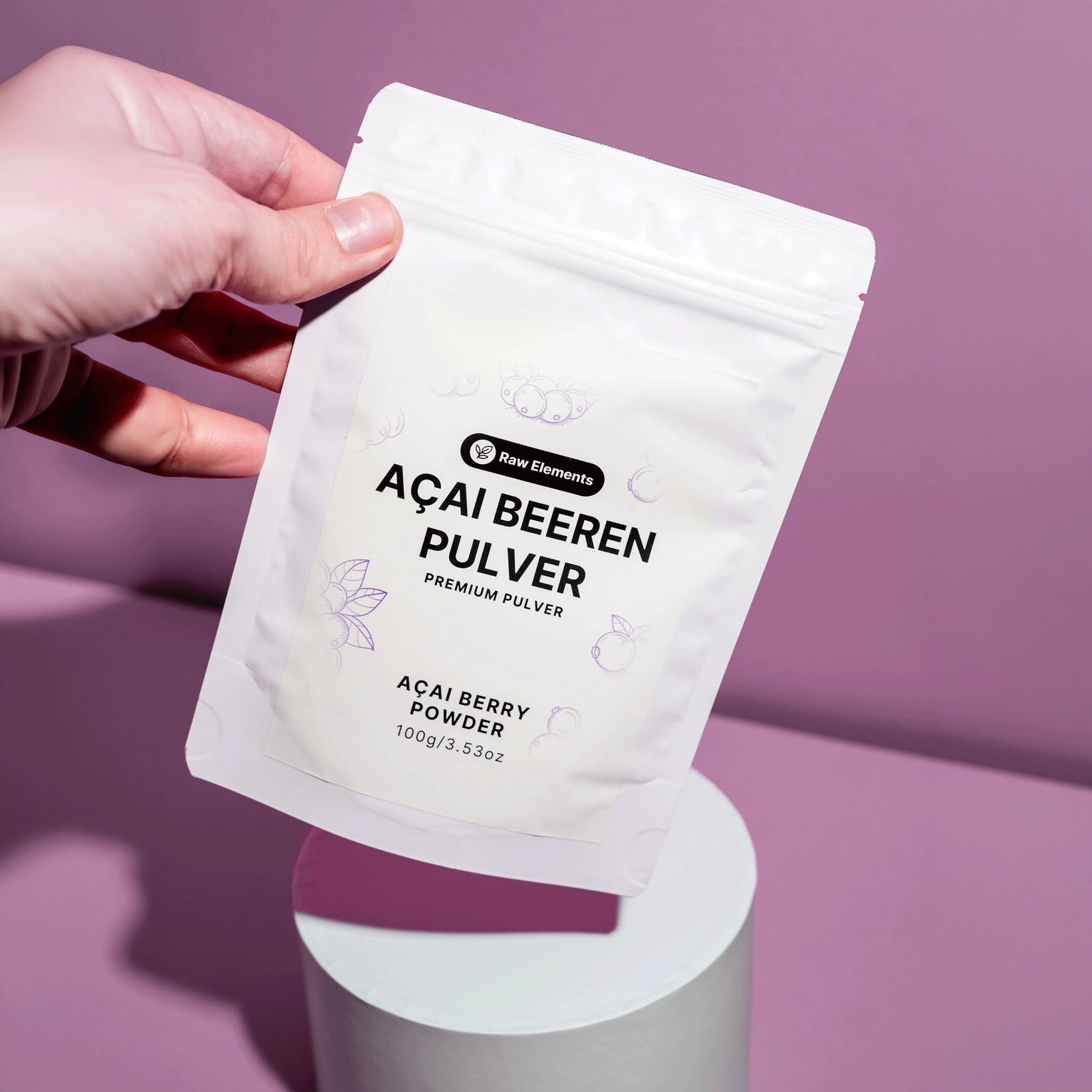 Eine Hand hält die Raw Elements Bio Açai Beeren Pulver Verpackung schwebend über einem weißen Podest.