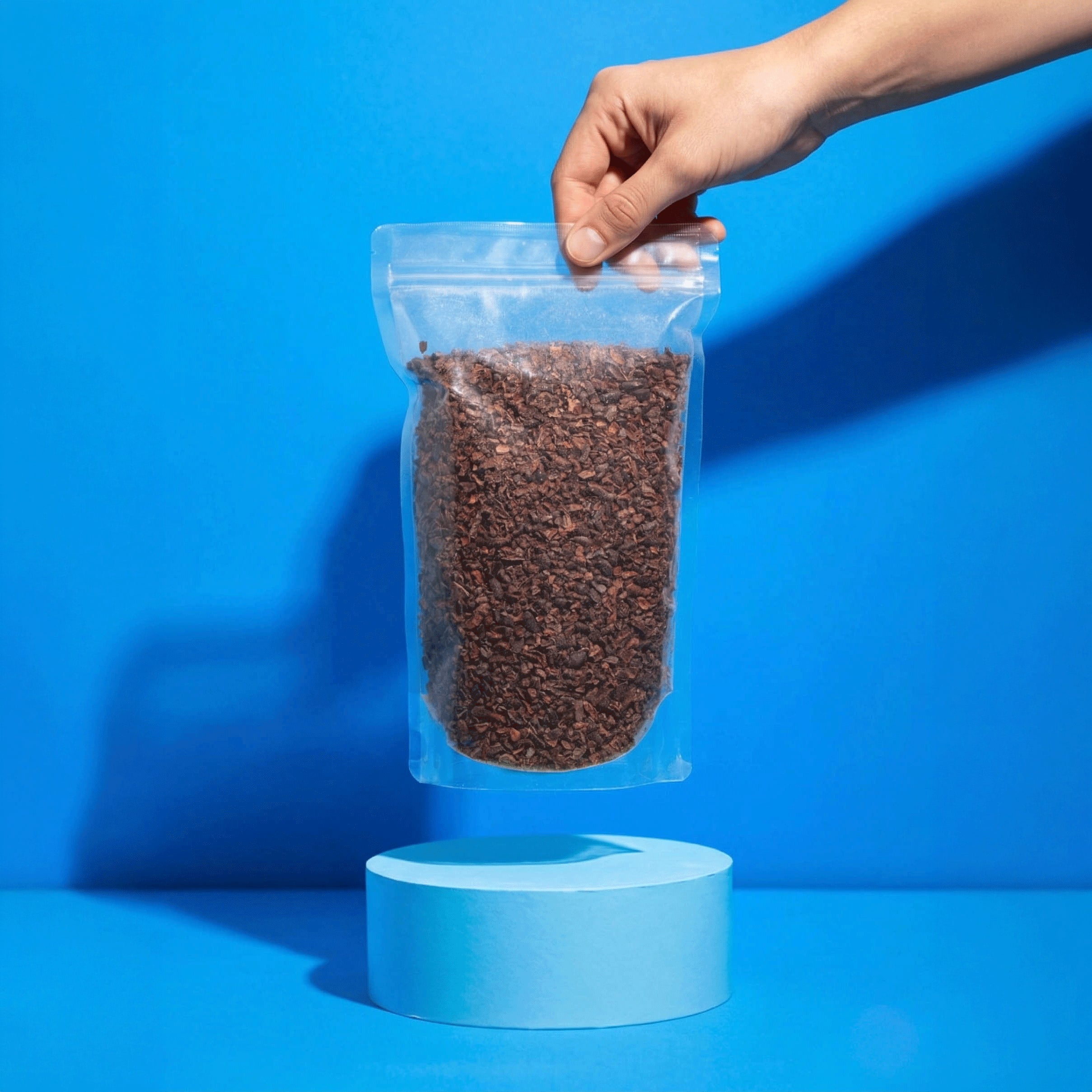 Eine Hand präsentiert einen transparenten Beutel mit Bio Kakaonibs schwebend über einem runden, hellblauen Podest.