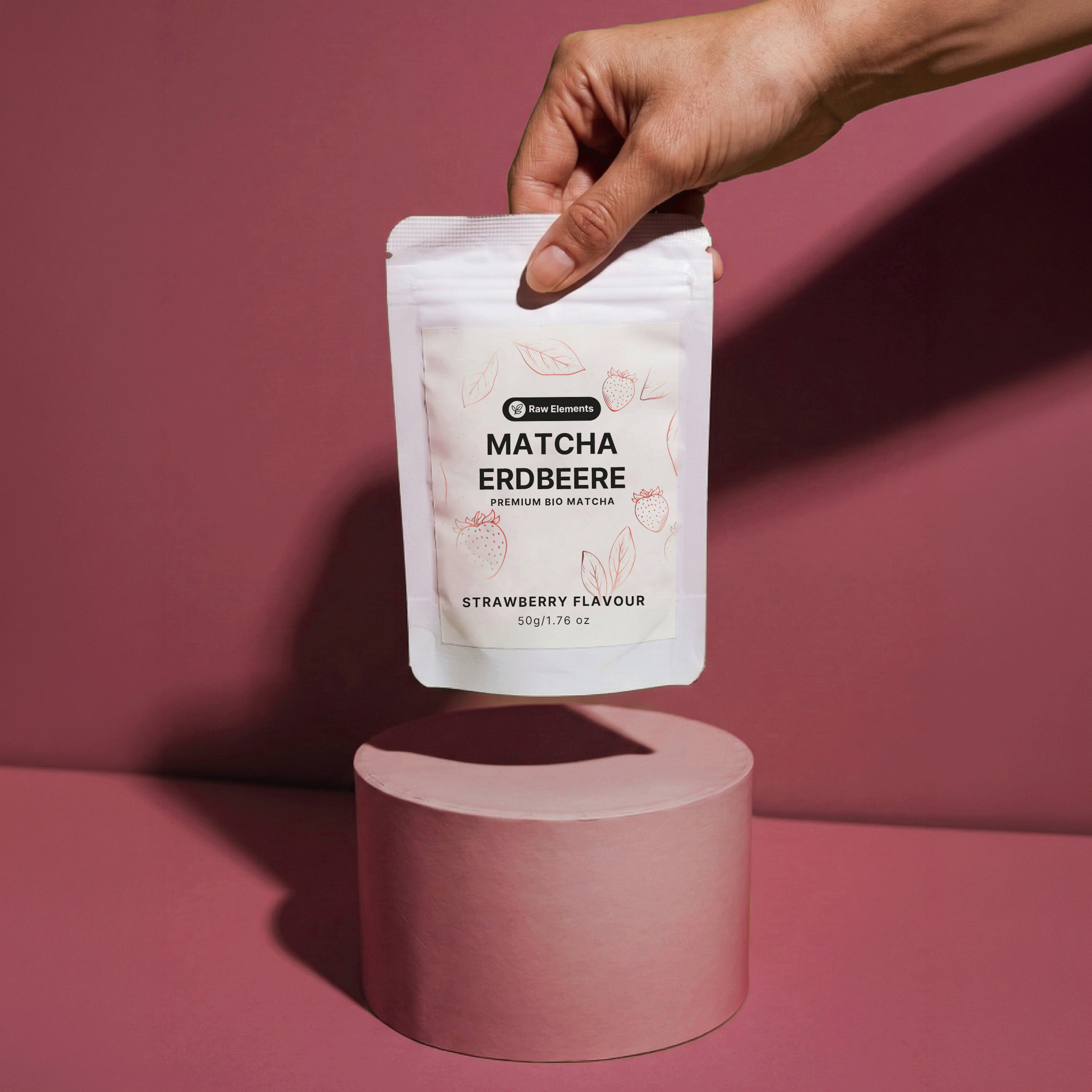 Eine Hand präsentiert die Raw Elements Bio Matcha Erdbeere Verpackung schwebend über einem altrosa Podest vor farblich passendem Hintergrund.