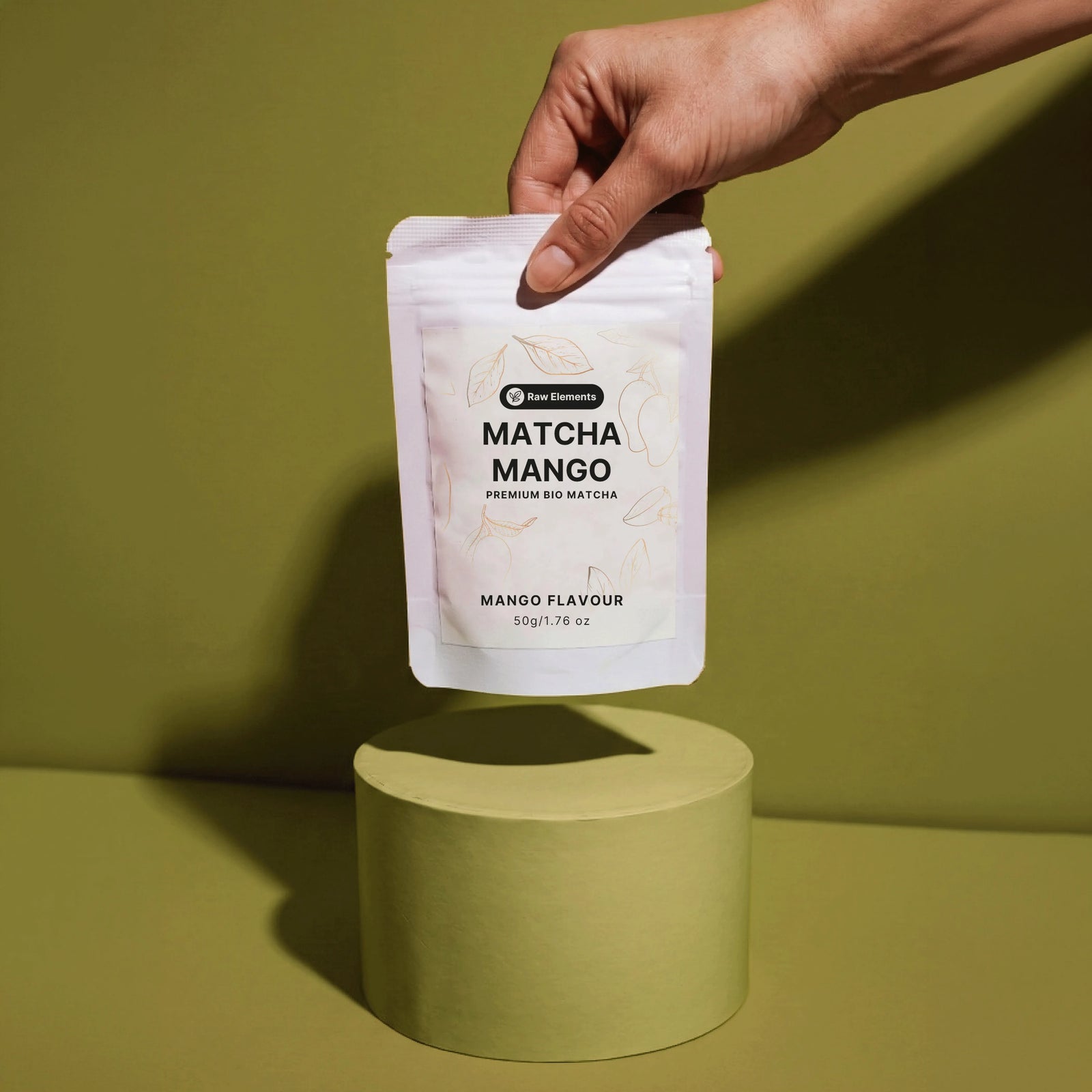 Eine Hand hält die Raw Elements Bio Matcha Mango Verpackung schwebend über einem hellgrünen Podest