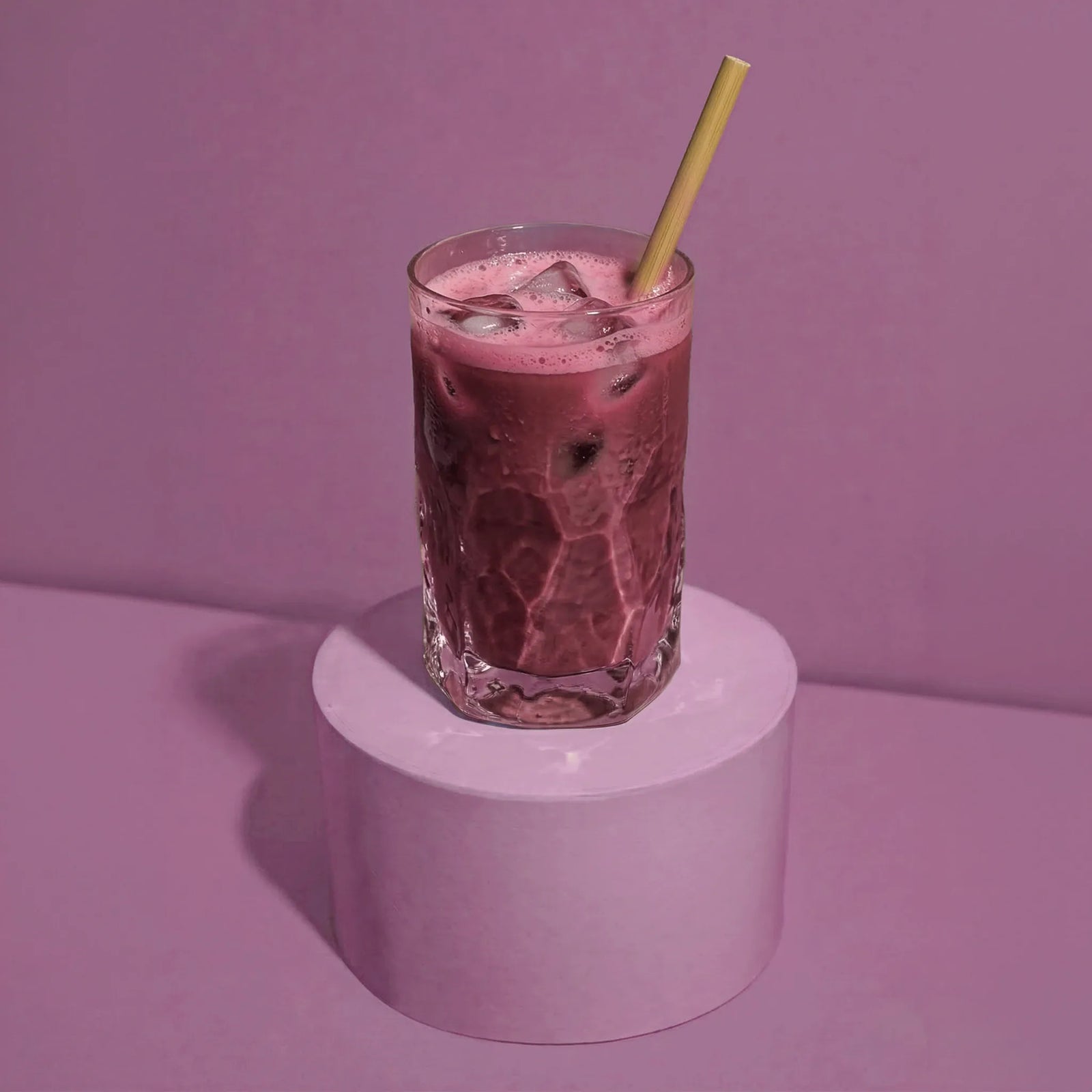 Erfrischender Bio Acai-Beeren Drink mit Eiswürfeln und Bambusstrohhalm im Glas auf violettem Podest