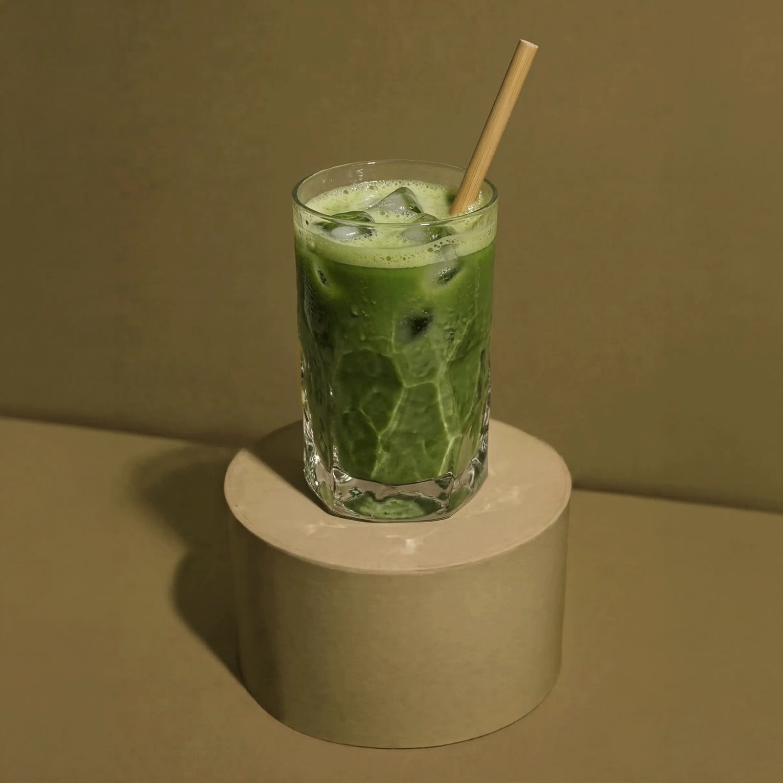 Ein erfrischender Bio-Hojicha Iced Latte mit Eiswürfeln und einem Bambusstrohhalm in einem Glas, stehend auf einem runden Podest.