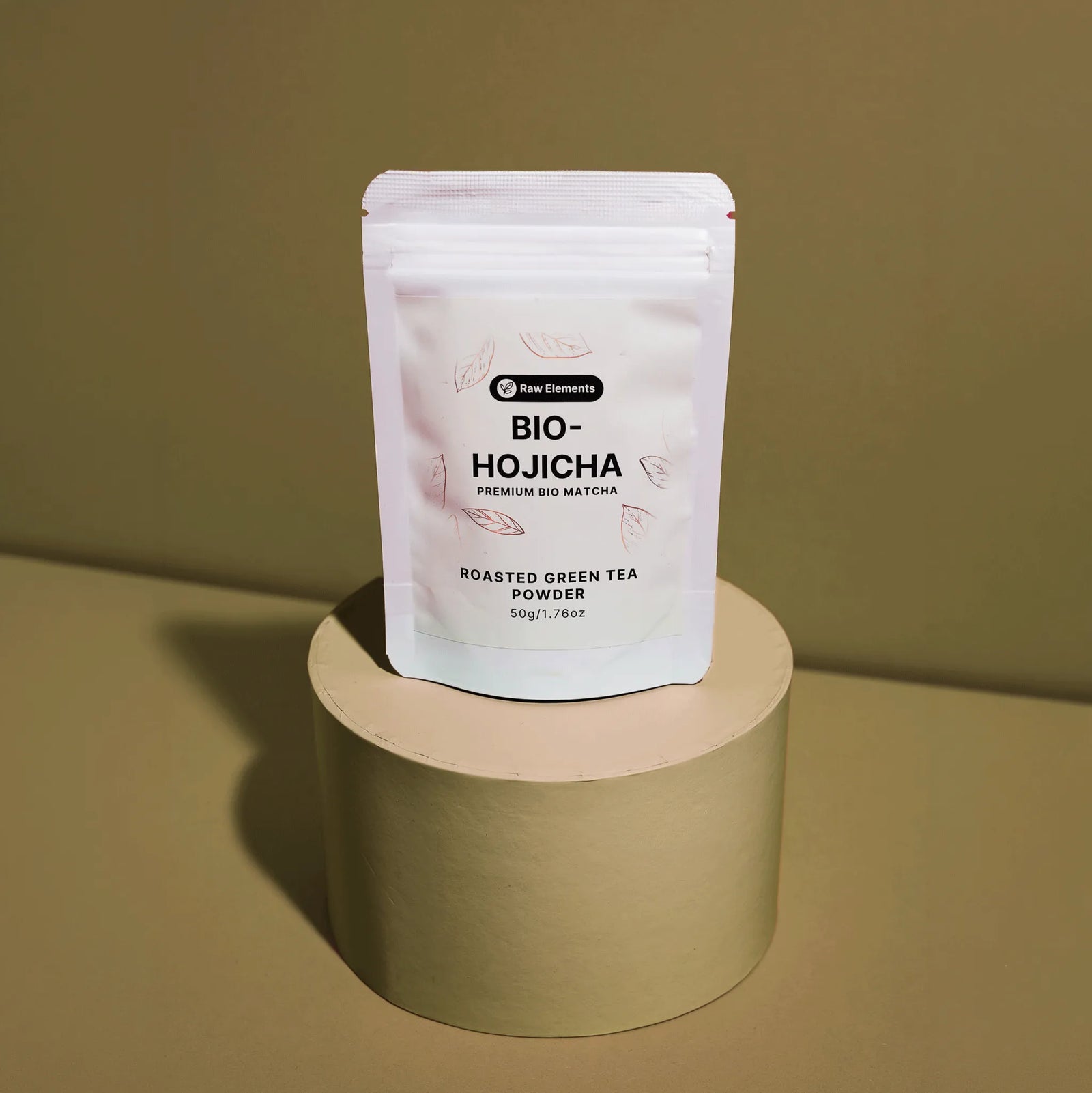 Weißer Standbodenbeutel des Raw Elements Bio-Hojicha (Roasted Green Tea Powder), platziert auf einem runden, erdigen Podest.