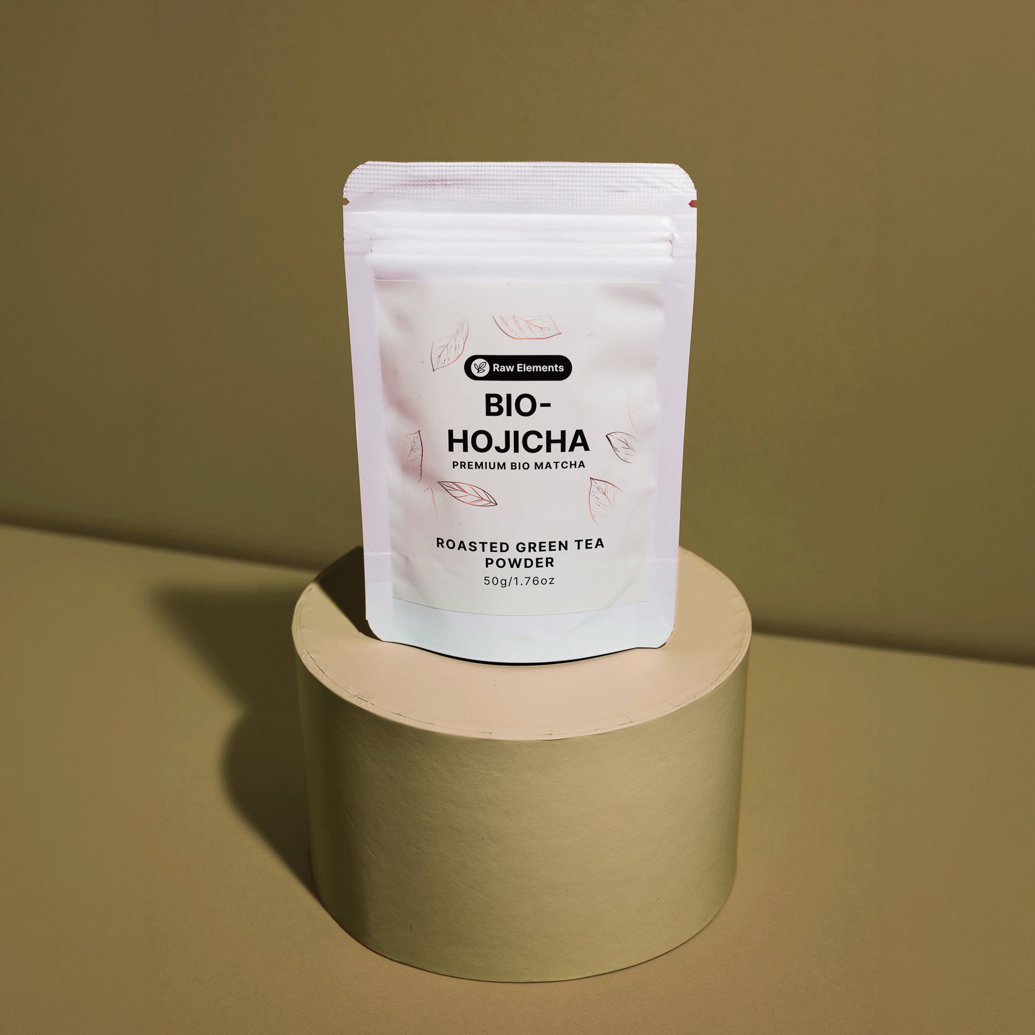 Weißer Standbodenbeutel des Raw Elements Bio-Hojicha (Roasted Green Tea Powder), platziert auf einem runden, erdigen Podest.