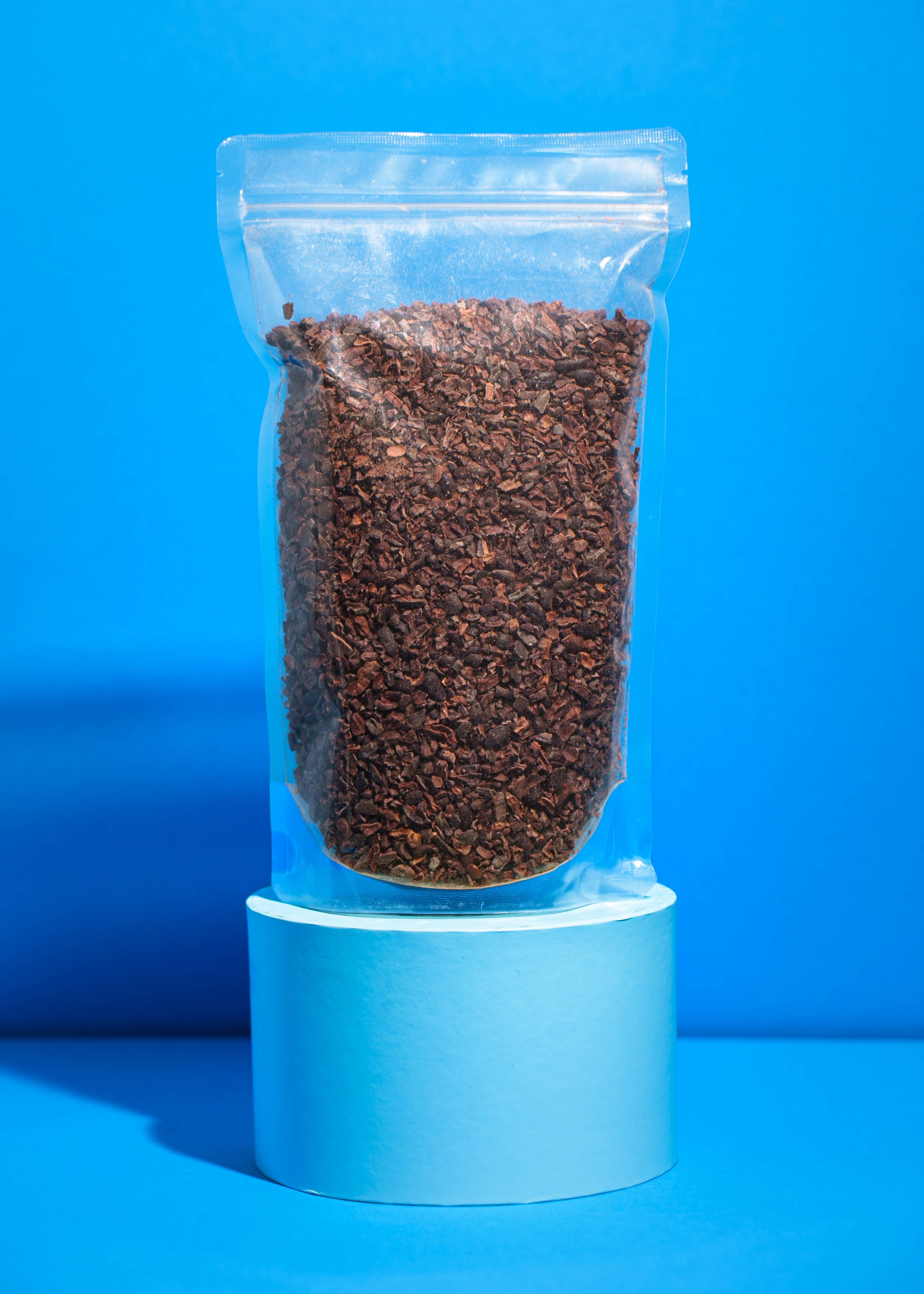 Transparente 500g Standbeutel-Packung mit Bio Kakaonibs aus Madagaskar vor blauem Hintergrund.