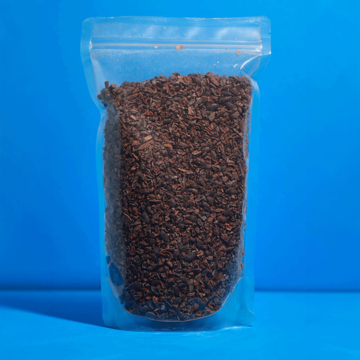 Transparenter Standbodenbeutel gefüllt mit 500g reinen Bio Kakaonibs auf blauem Hintergrund.