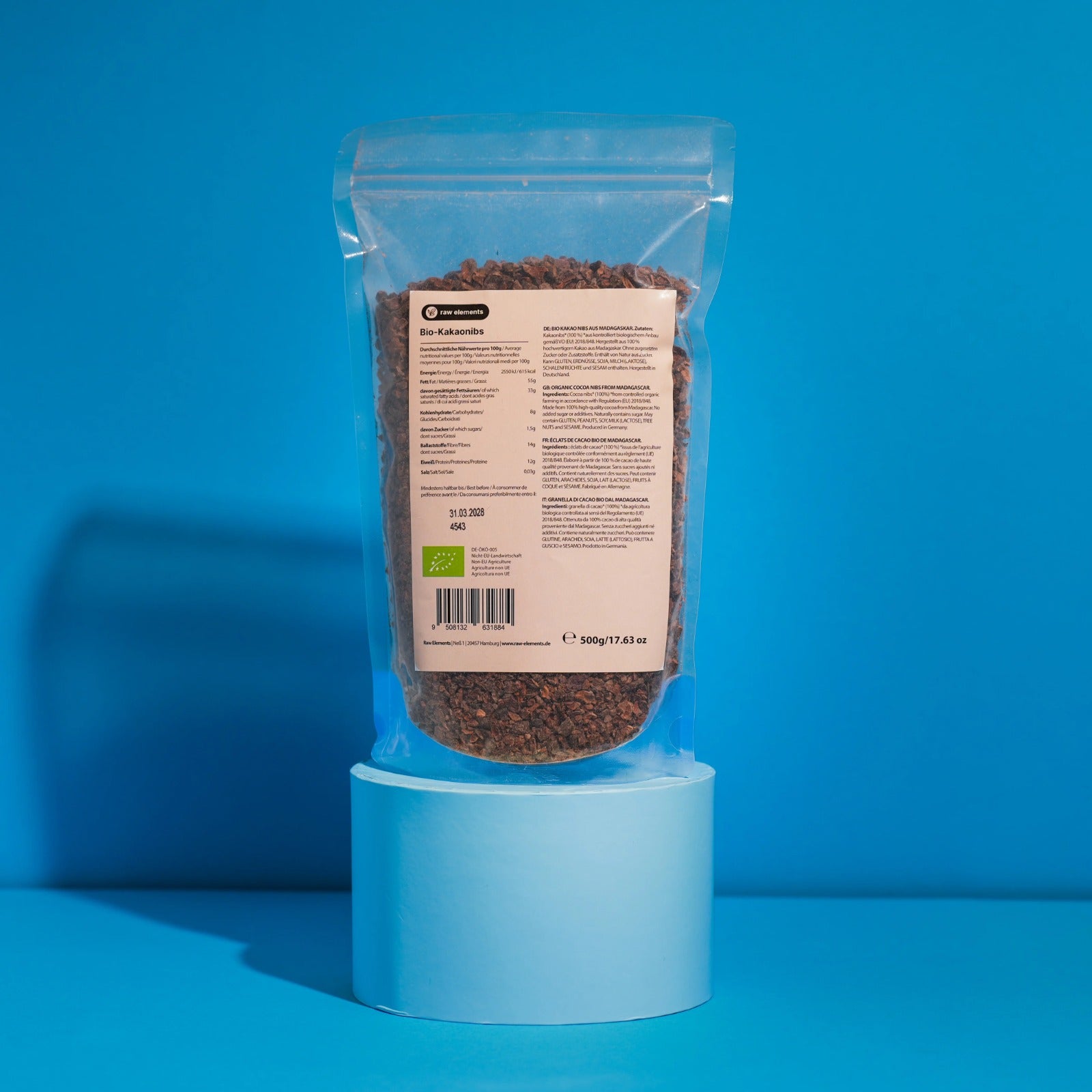 Transparenter Standbodenbeutel mit 500g reinen Bio Kakaonibs auf einem weißen Podest vor blauem Hintergrund. Ansicht der Rückseite mit Etikett.