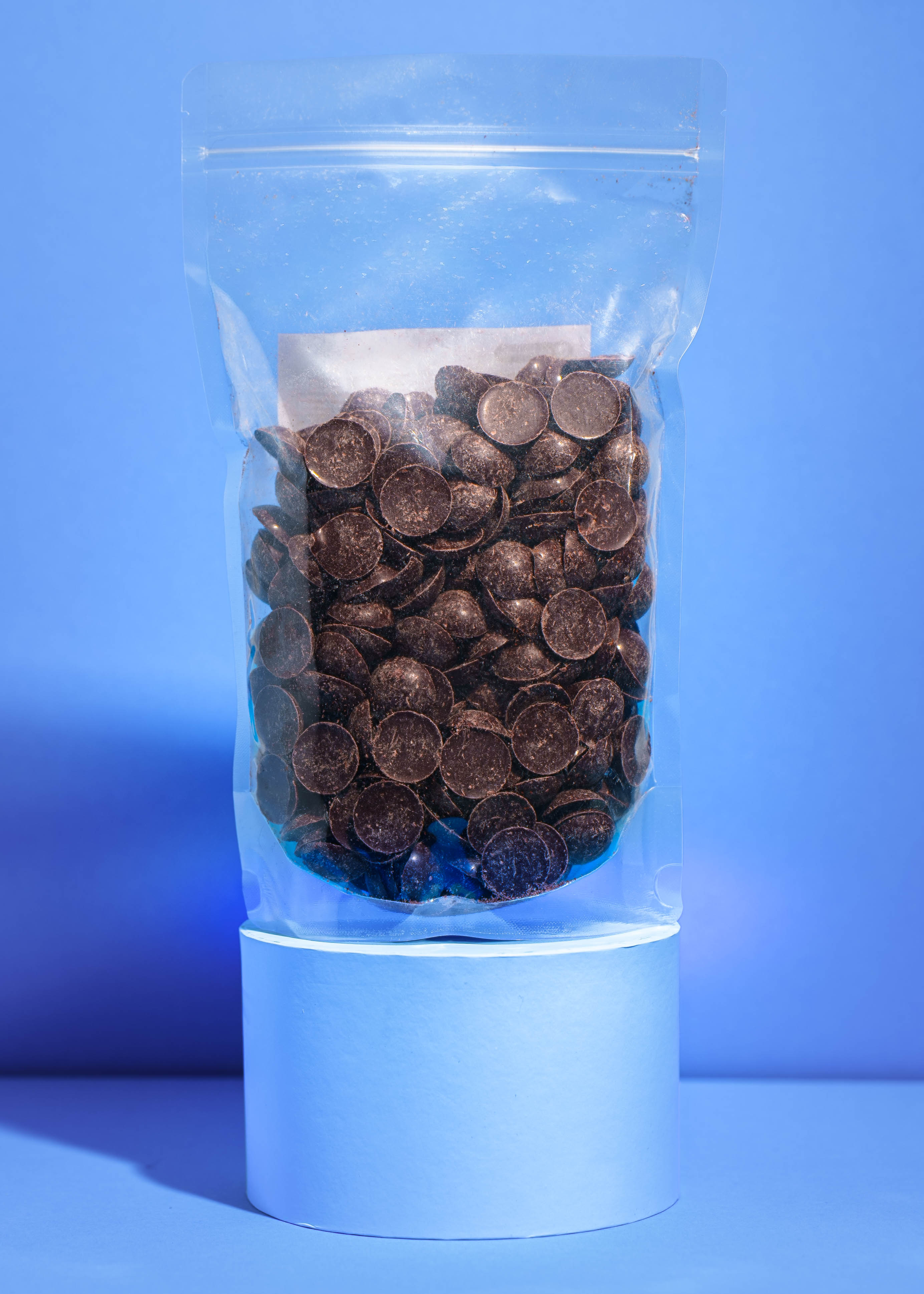 Der 1kg-Beutel mit Bio-Schokoladendrops, nur mit Dattelsüße gesüßt, präsentiert auf einem hellblauen runden Podest vor einem blauen Hintergrund.