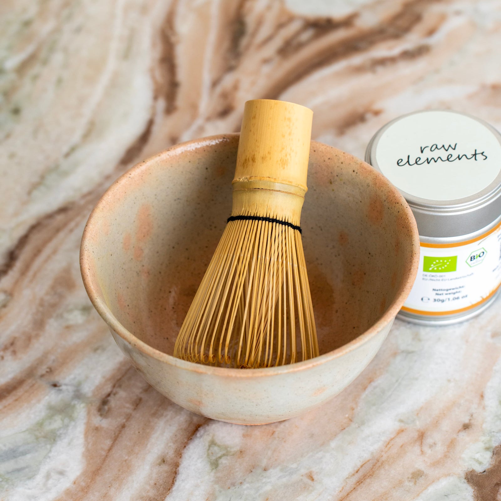 Chasen Matchabesen aus Bambus in einer beigen Matchaschale neben einer Raw Elements Bio Matcha Dose