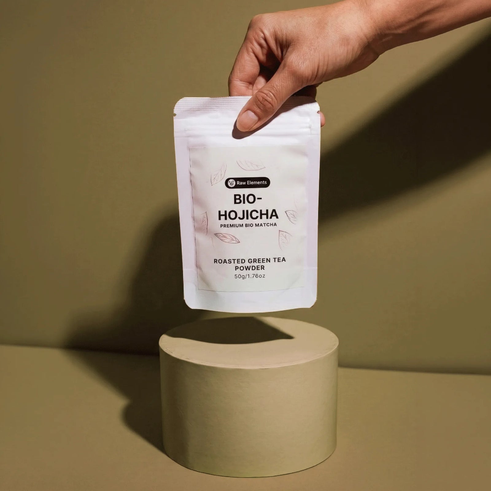Eine Hand hält die weiße Raw Elements Bio-Hojicha Verpackung schwebend über einem Podest vor erdigem Hintergrund.