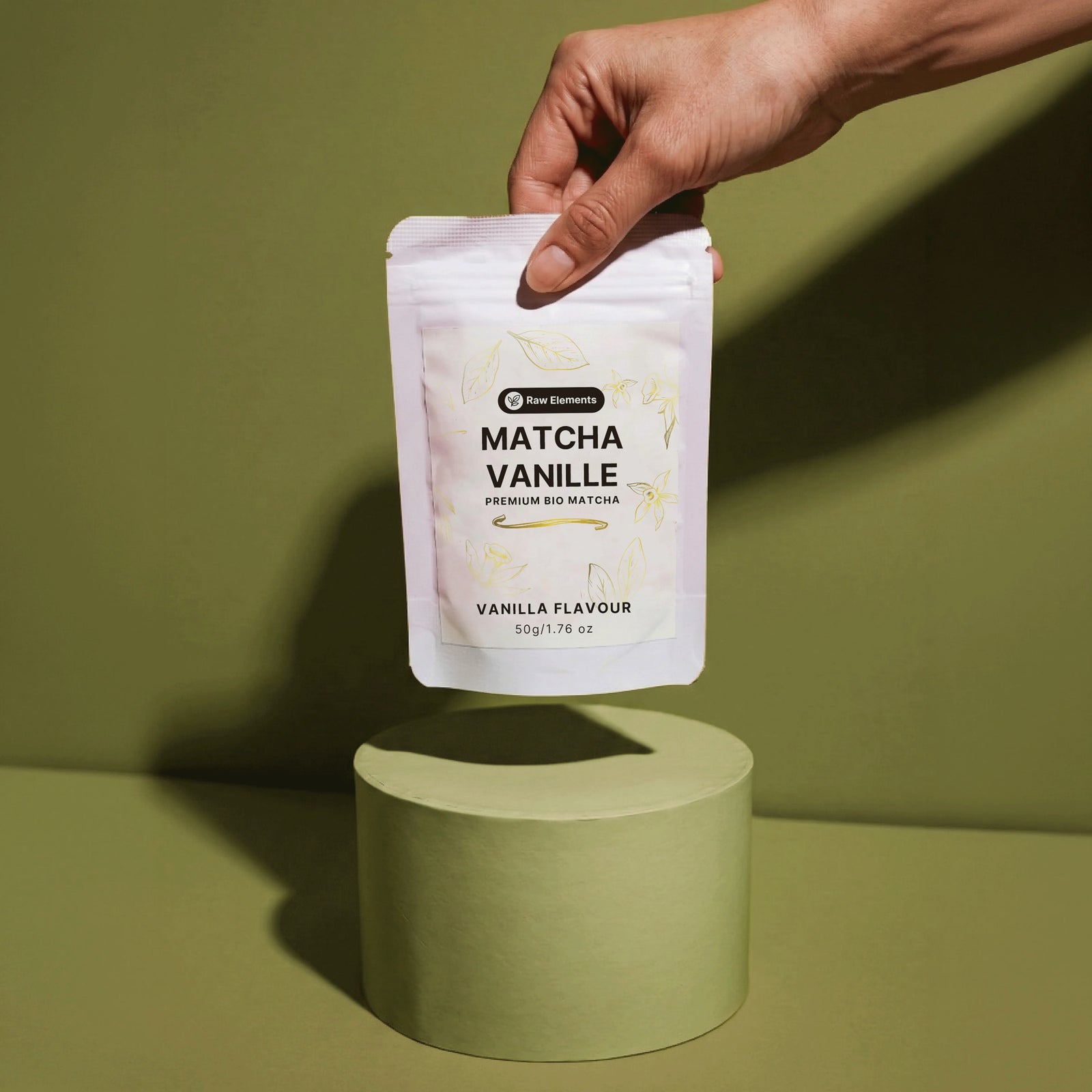 Eine Hand präsentiert die Raw Elements Bio Matcha Vanille Verpackung schwebend über einem grünen Podest.