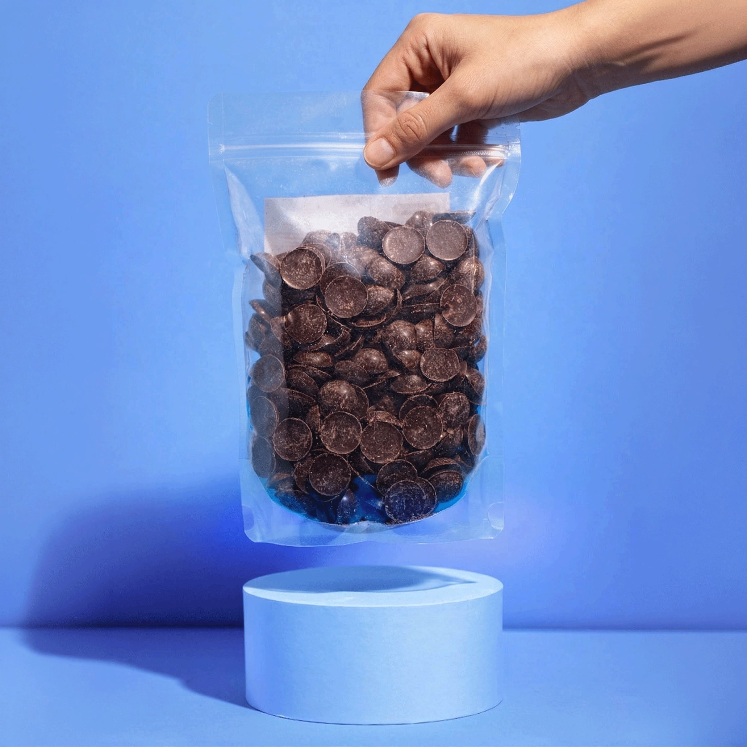 Eine Hand hält einen transparenten Beutel mit Bio Schokodrops schwebend über einem hellblauen Podest.