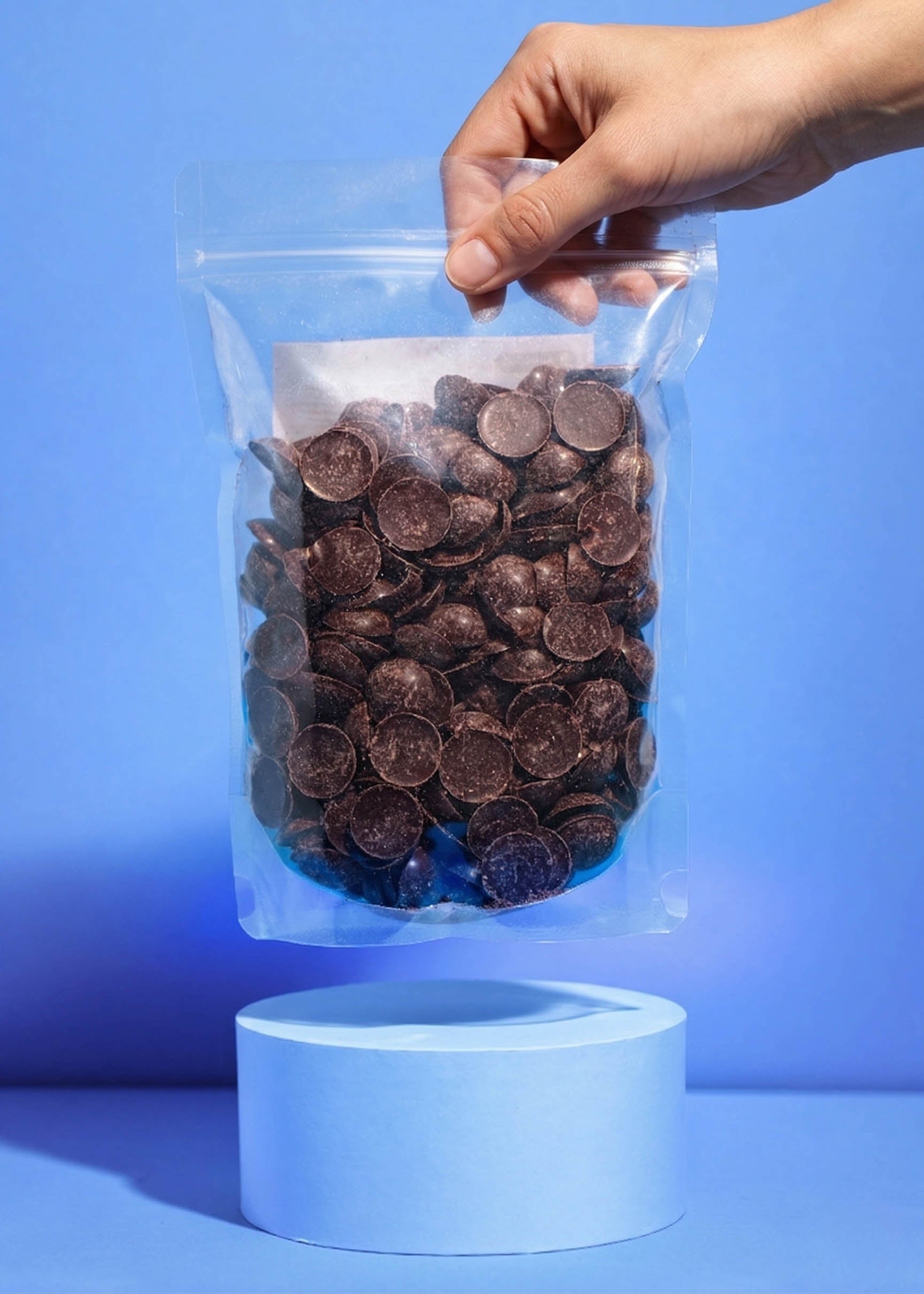 Eine Hand hält den schwebenden 1kg-Beutel der Raw Elements Bio-Schokoladendrops mit Dattelsüße über einem hellblauen Podest. Der transparente Beutel zeigt die dunklen Schokodrops