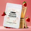 Weiße Pouch-Verpackung von Raw Elements Premium Matcha Erdbeere, umgeben von frischen Erdbeeren und einem traditionellen Bambusbesen.