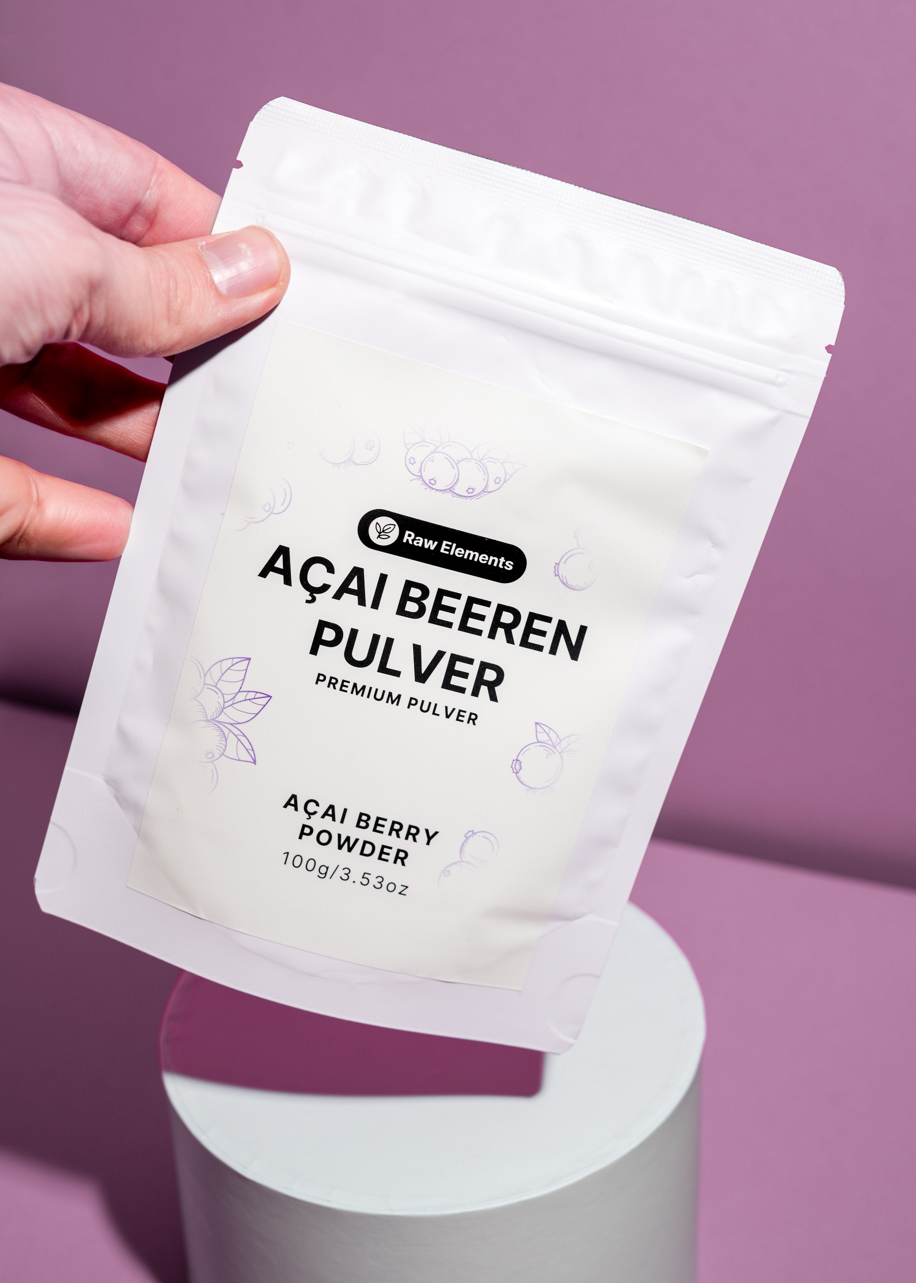 Eine Hand hält den weißen Beutel mit Acai Matcha Pulver schräg über ein weißes Podest vor lila Hintergrund